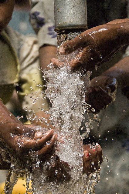 Multiple pairs of hands reaching under a flowing water pipe, sharing a scarce communal water source